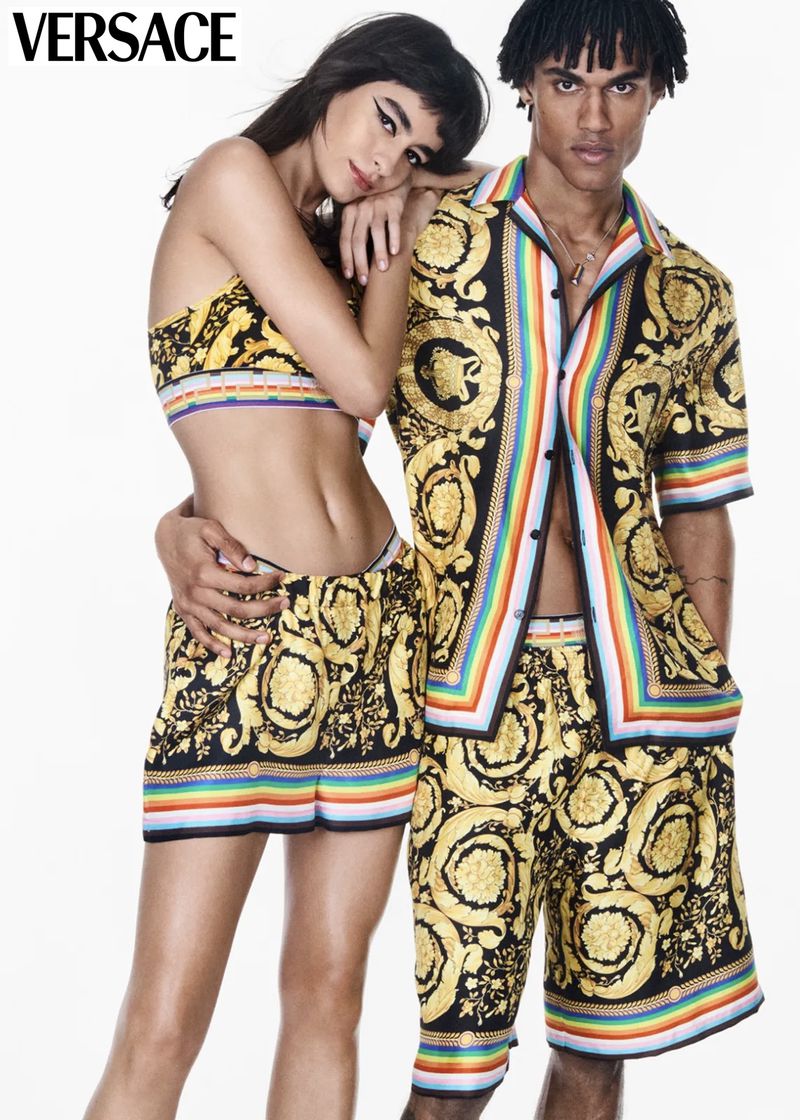 Two fashion models pose in matching Versace-print black and gold shorts and tops with rainbow detail against a white backdrop in a studio shot.