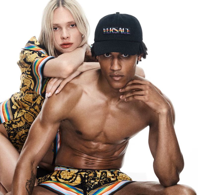 Fashion shot of a man with a Versace cap and printed shorts alongside a woman wearing a patterned shirt against a white backdrop.