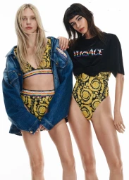 Two models posing in Versace gold and black baroque print swimwear with rainbow Pride details against a white seamless background.