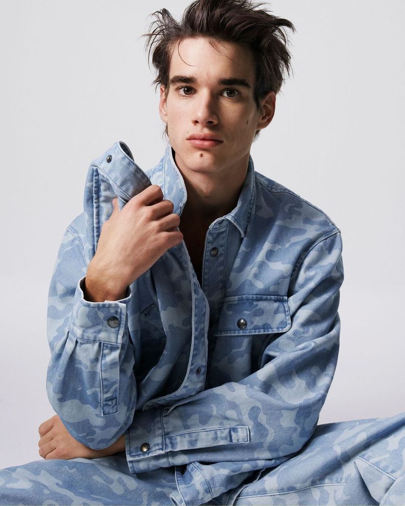 An adult male in a light-blue camo denim jacket and pants sits in a photo studio with a soft white background and diffused light.
