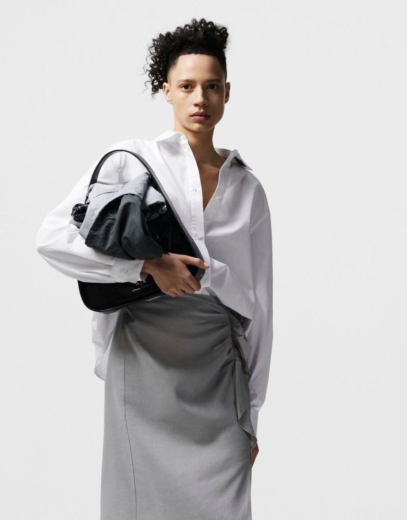 Adult fashion model poses in a photo studio, holding a black handbag while wearing a long gray skirt and a white button-down shirt.