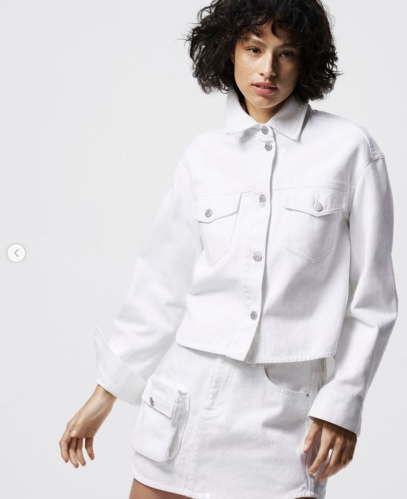 A woman poses in a short white denim skirt and matching jacket against a neutral backdrop, showcasing a fashion-forward, monochromatic look.