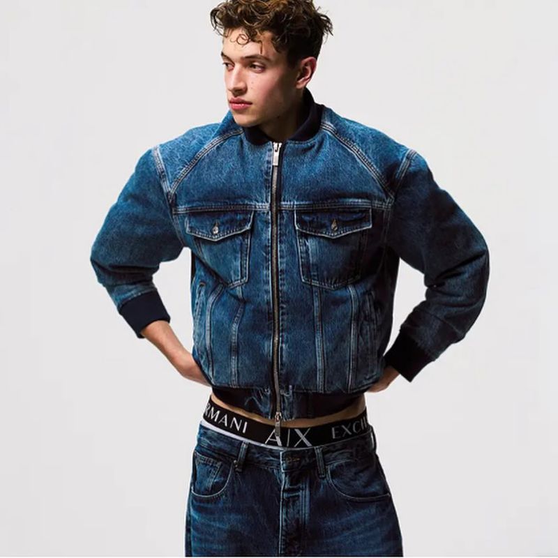 Male fashion model poses in a studio wearing a denim jacket and jeans, showcasing a casual yet stylish look against a neutral backdrop.