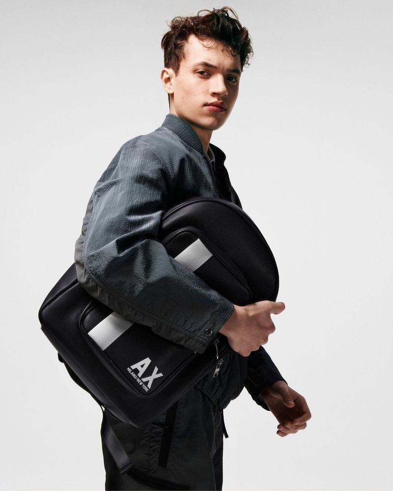 Young adult male model in grey jacket holds black AX Armani Exchange laptop bag in a studio shot against a gradient background.