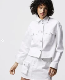 Model wearing a white buttoned denim jacket and matching white mini skirt against a bright studio background.