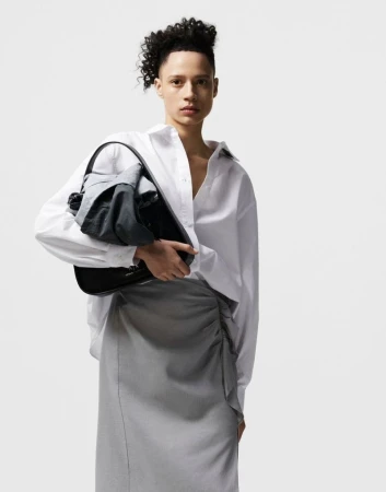 Model holding a black handbag with wrapped grey fabric, wearing a crisp white button-down shirt and grey skirt against a white studio sweep.