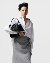 Model holding a black handbag with wrapped grey fabric, wearing a crisp white button-down shirt and grey skirt against a white studio sweep.