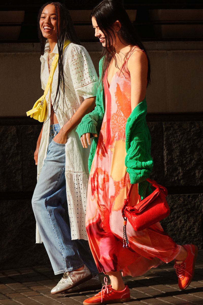 Two women stroll outdoors, showcasing vibrant summer fashion. One wears a floral maxi dress and green cardigan, the other a white eyelet dress and jeans.