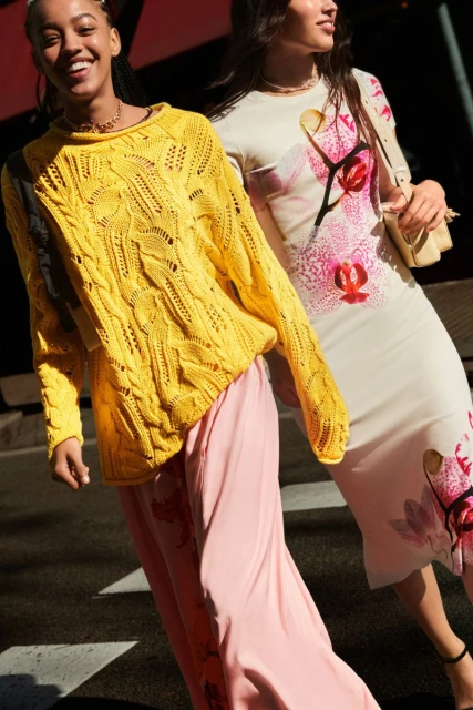 Two women walk on a crosswalk wearing fashionable clothing. One is wearing a yellow knitted sweater and the other a floral dress.
