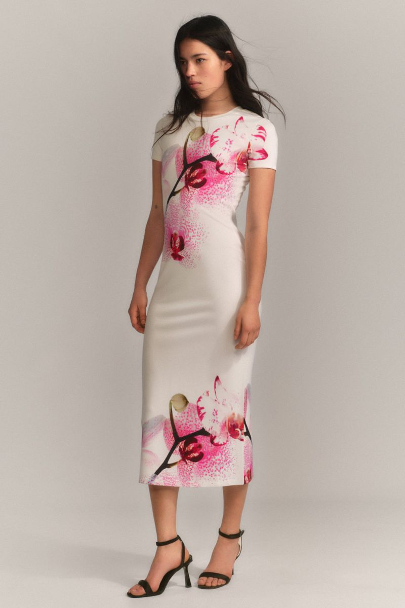 A woman in a cream dress with pink flower print and black high heels stands in a photo studio with a light background.