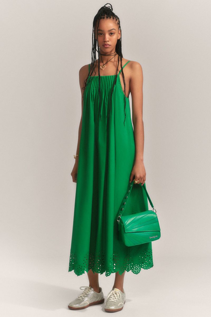 A biracial fashion model poses in a green dress holding a matching handbag, in a studio setting. She wears gold sneakers.