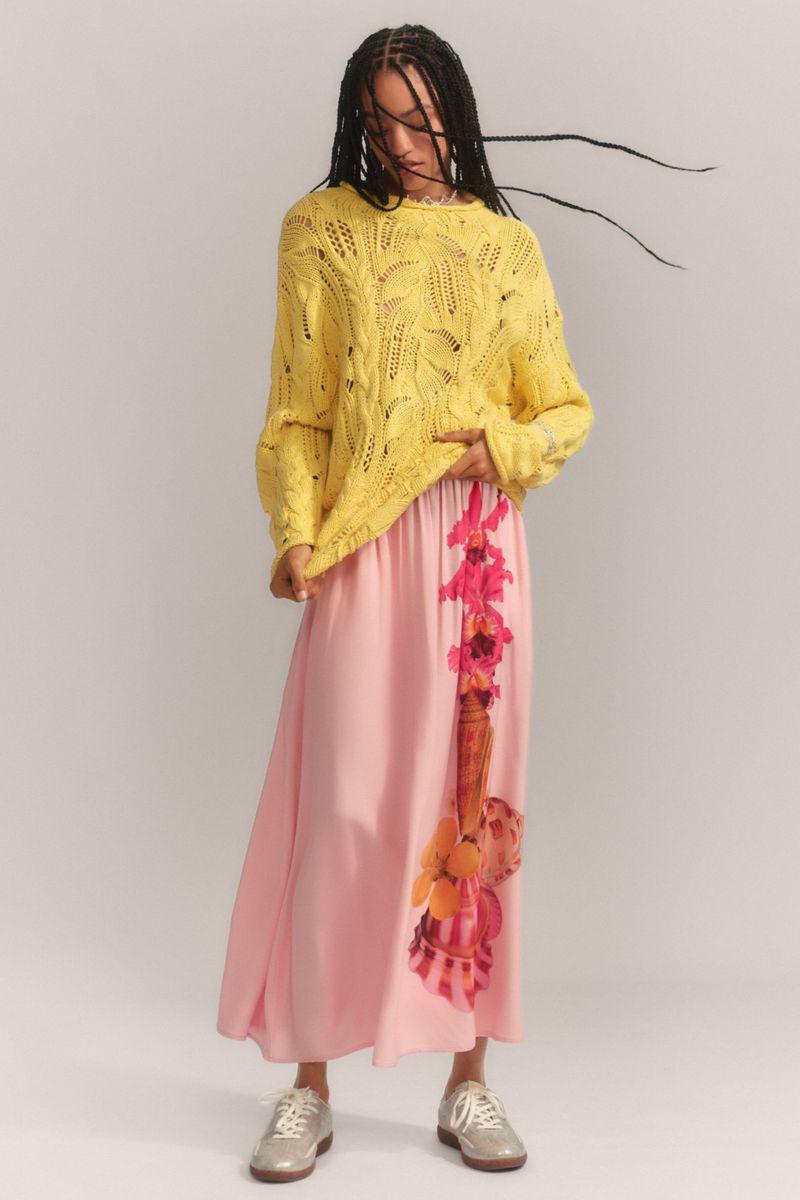 A woman poses in a yellow sweater and pink floral-print skirt against a white background in a full shot with soft studio lighting.