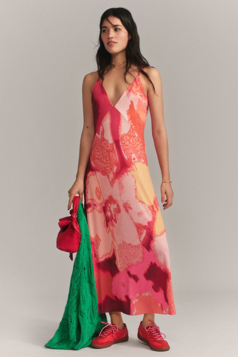 Brunette woman posing in a pink patterned dress, holding a red purse and green knit shawl against a neutral backdrop.