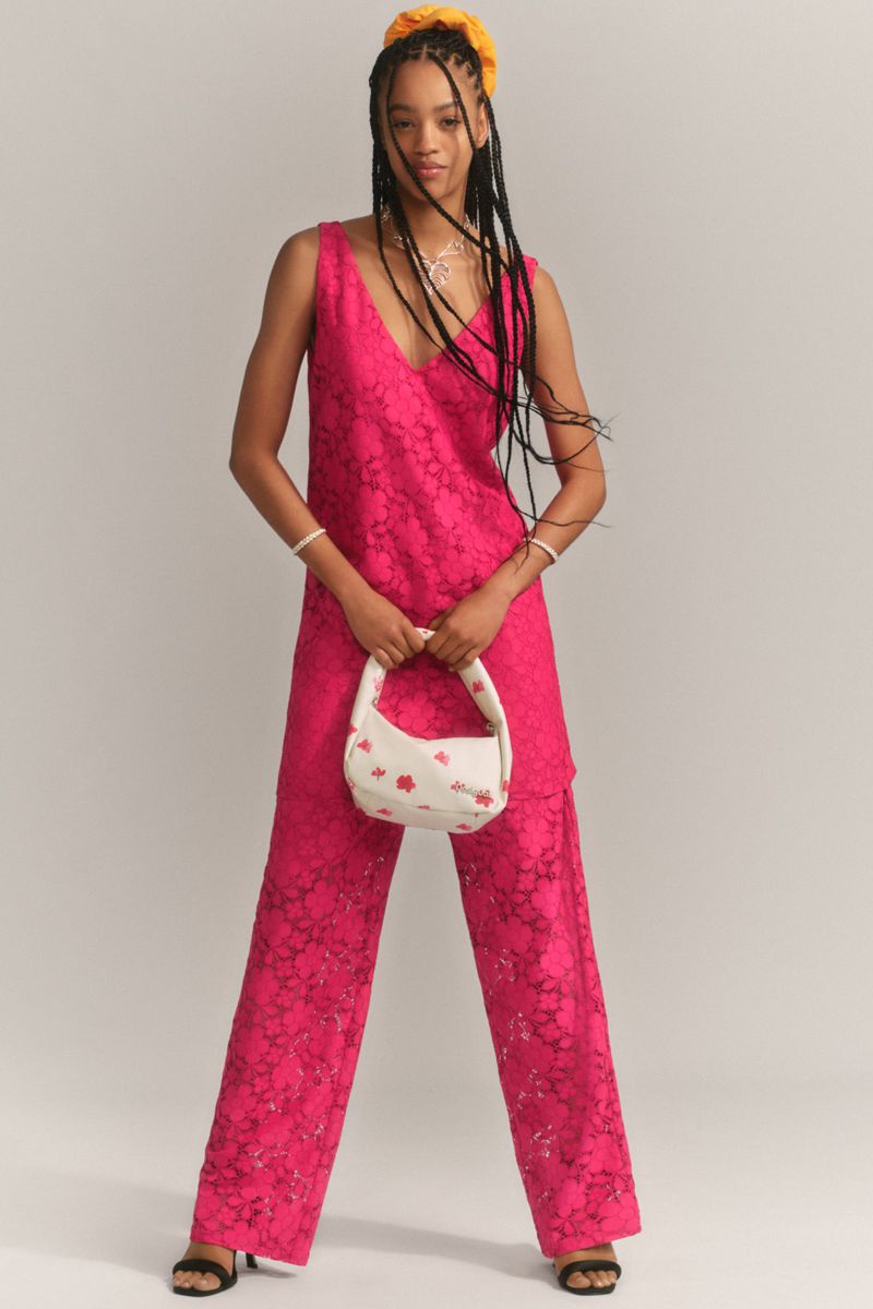 A slender woman with braids stands in a photo studio, modeling a bright-pink lacy jumpsuit with a small white purse and black heeled shoes.