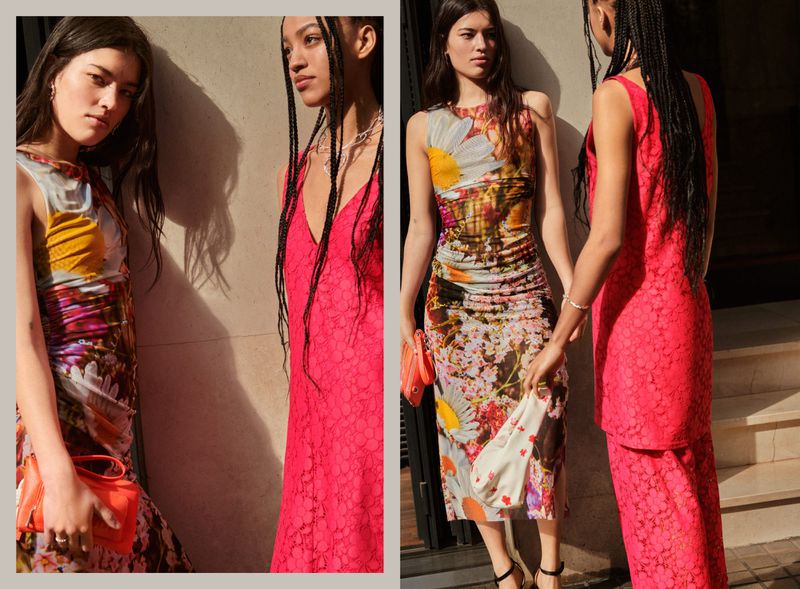Two women in floral dresses pose outdoors. One woman wears a pink dress, while the other carries an orange clutch in natural lighting.