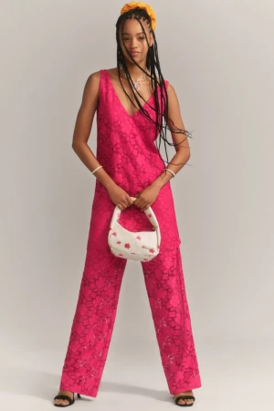 Model wearing a bright pink lace tunic and matching trousers while holding a white embroidered handbag in a studio.