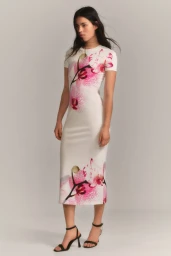 Model posing in a white short-sleeve midi dress featuring pink orchid floral prints and black strap heels against a studio backdrop.