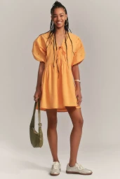 Smiling woman with long braids posing in an orange puff-sleeve mini dress holding an olive green bag against a grey studio backdrop.