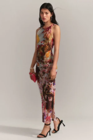 Model posing in a floral print sleeveless maxi dress with strappy black heeled sandals and a pink clutch against a grey studio backdrop.