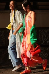 Two women walk together in bright sunshine wearing vibrant summer dresses, a green cardigan, wide-leg jeans, and colourful bags.