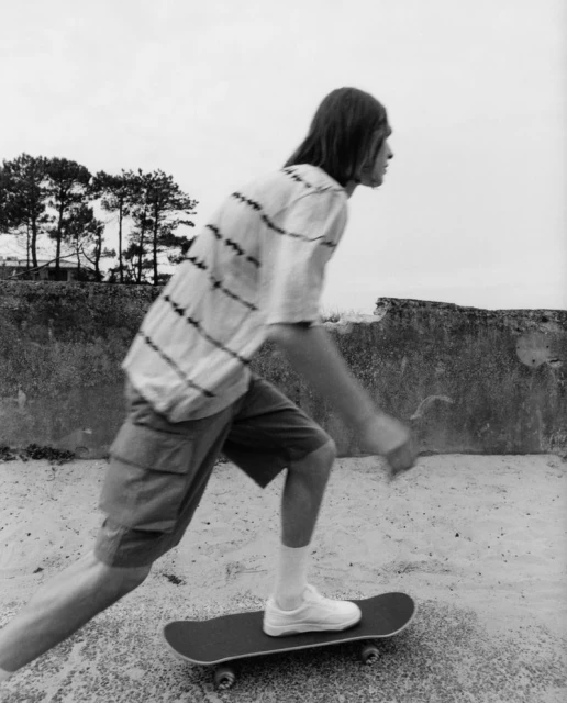 Black and white photograph of a young adult riding a skateboard in an outdoor environment with trees and a wall in the background.