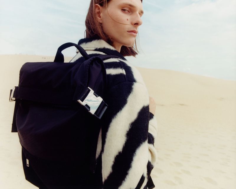 Person with long hair and a zebra-striped jacket poses with a black backpack against a background of sand dunes and a light blue sky.