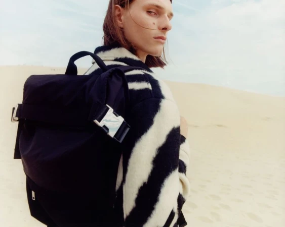 Person with long hair and a zebra-striped jacket poses with a black backpack against a background of sand dunes and a light blue sky.