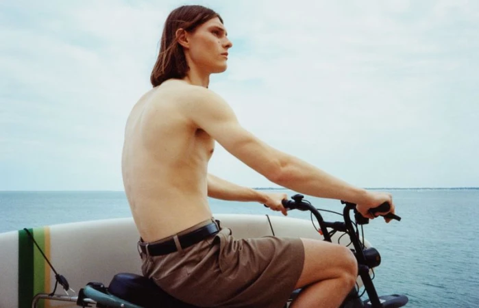 A shirtless man with long brown hair rides a motorized bicycle near the ocean, surfboard attached to the side, under a cloudy sky.