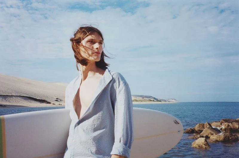 A long-haired man in a light blue shirt holds a surfboard on a beach with sand dunes and ocean under a blue sky with white clouds.