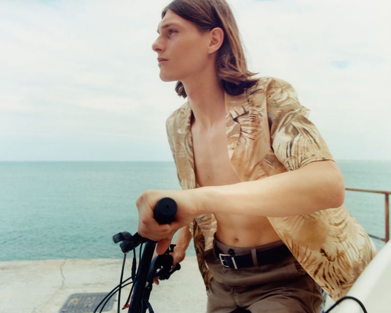 Young man with long hair stands with a bicycle overlooking the ocean on a sunny day, wearing a patterned shirt and brown shorts.