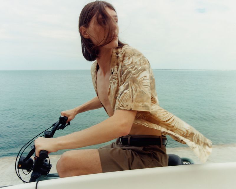 A person riding a bicycle along a concrete path with the ocean in the background, wearing a patterned shirt and shorts.