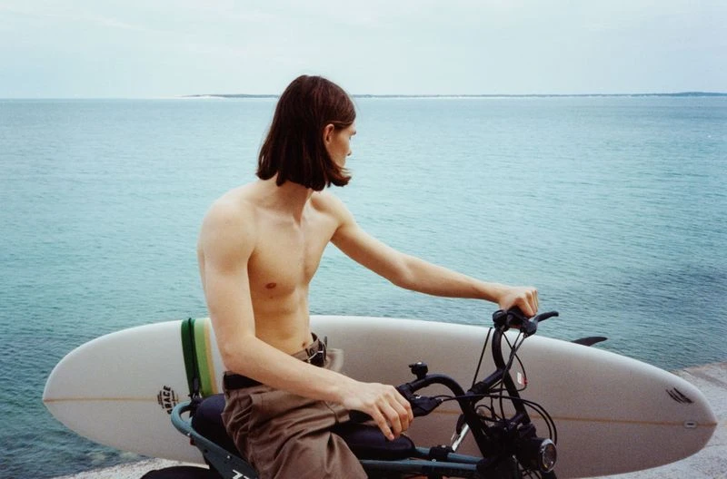 Shirtless man on electric bicycle with surfboard at the beach with ocean and horizon background. Lifestyle and recreation visual.