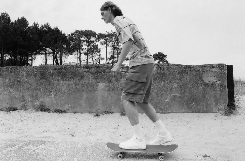 Black and white outdoor shot of a young adult riding a skateboard on a concrete path, wearing shorts and a striped t-shirt.