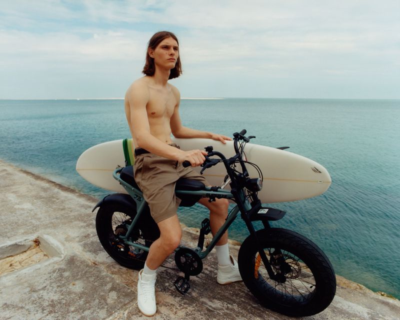 Shirtless man with surfboard rides electric bicycle by blue ocean against cloudy sky; sporty lifestyle, outdoor recreation, and sustainable transport.
