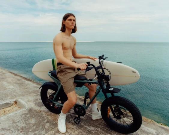 Shirtless man with surfboard rides electric bicycle by blue ocean against cloudy sky; sporty lifestyle, outdoor recreation, and sustainable transport.