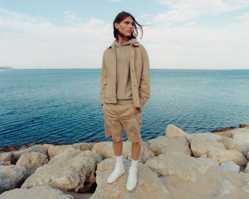 A young adult male poses on rocks near a body of water wearing a beige hooded sweatshirt, jacket, shorts, and white shoes on a sunny day.