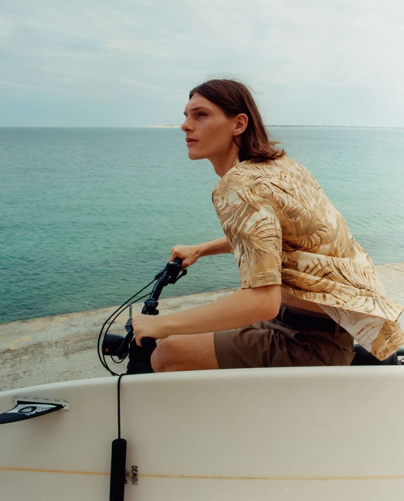 A young man on a bicycle carrying a surfboard, riding near the ocean on a sunny day, wearing a patterned shirt and brown shorts.