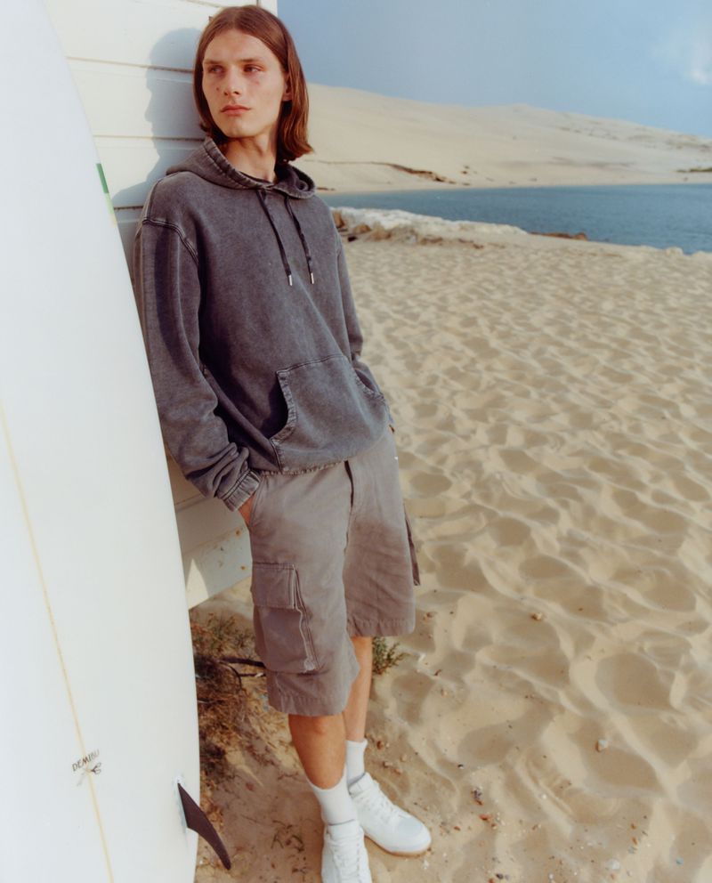 Man in hoodie and shorts stands next to surfboard on beach near water and sand dune.
