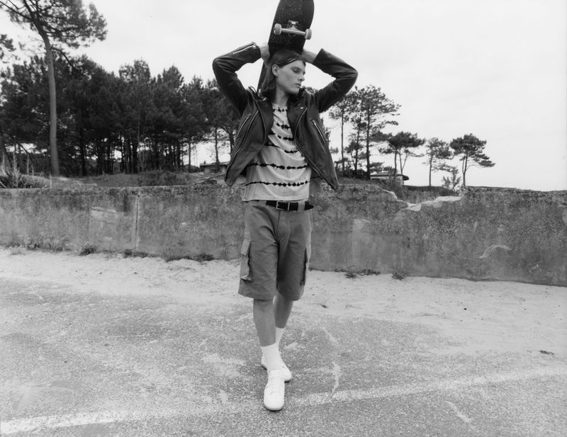 Monochrome portrait of a male model walking with a skateboard above his head, wearing casual street style attire, outdoors.