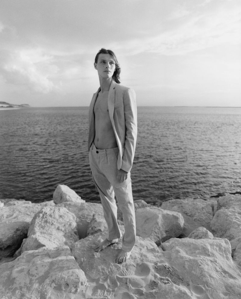 Black and white portrait of a young adult man in a suit jacket and pants standing on rocks by the sea under a cloudy sky.
