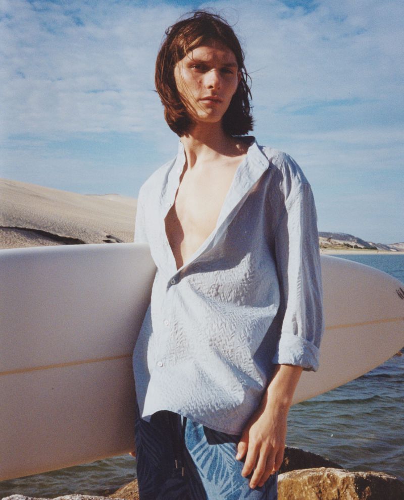 A fair-skinned adult male stands on a beach with a surfboard, wearing a light-blue patterned shirt and blue jeans with a palm leaf pattern.