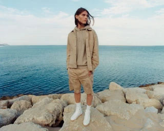 Model standing on coastal rocks wearing a beige jacket, hoodie, matching shorts, and white sneakers against an ocean background.