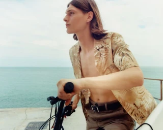 Teo Fortin wearing an unbuttoned printed shirt while holding bicycle handlebars against an open ocean backdrop.