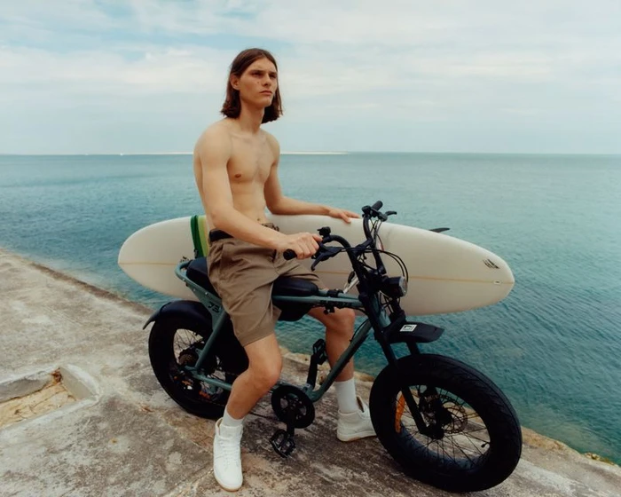 Shirtless model Teo Fortin sits on an electric bike with a mounted surfboard by the sea for the IRO S/S 24 lookbook.
