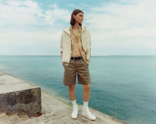 Model Teo Fortin in a cream knit cardigan, open patterned shirt, khaki shorts, and white sneakers on a sea breakwater.