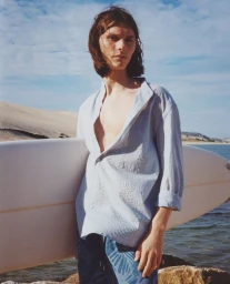 Model Teo Fortin holding a white surfboard wearing an unbuttoned light blue shirt in front of coastal sand dunes.
