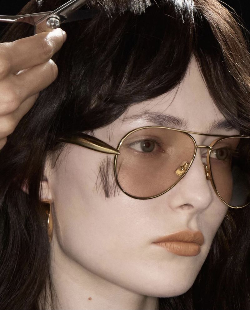 Close-up of a brunette adult woman with glasses having her bangs trimmed with silver scissors by a person out of frame.