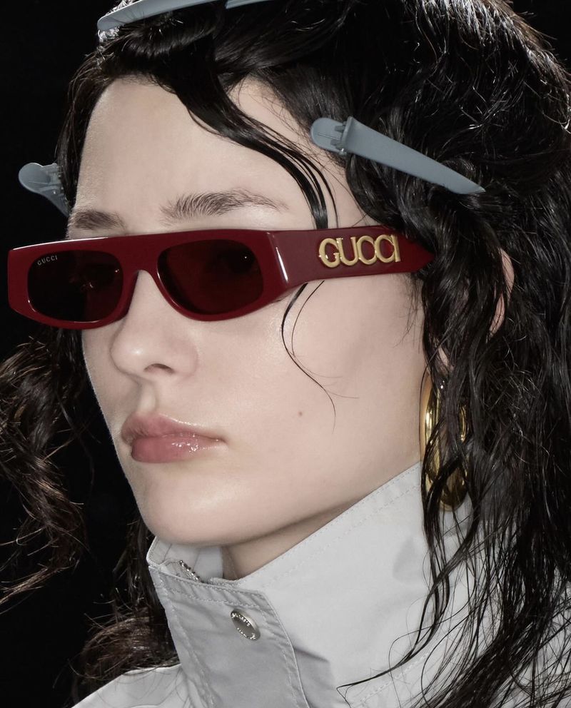 Close-up of model wearing burgundy Gucci rectangular sunglasses with gold branding and grey hair clips against a black background.