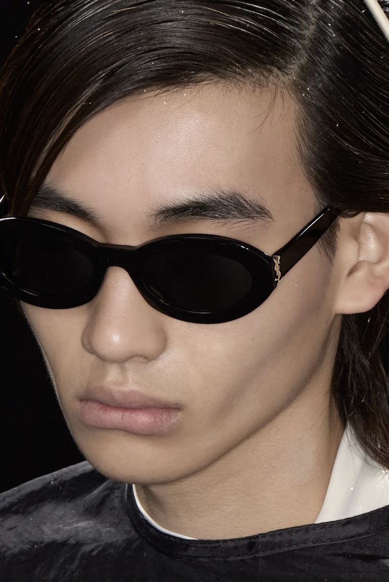 Close-up studio shot of a young adult male model wearing sunglasses, styled hair, and a dark shirt against a dark backdrop.