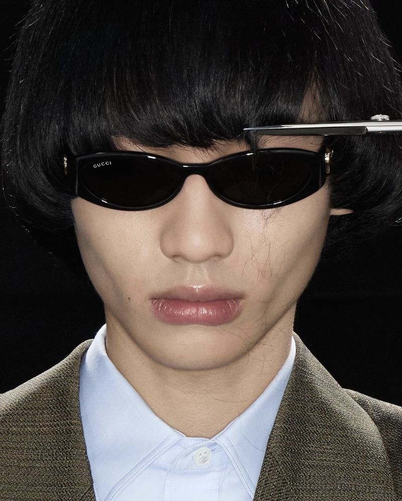 Close-up portrait of an East Asian model wearing black oval sunglasses getting bangs trimmed with scissors against a black background.